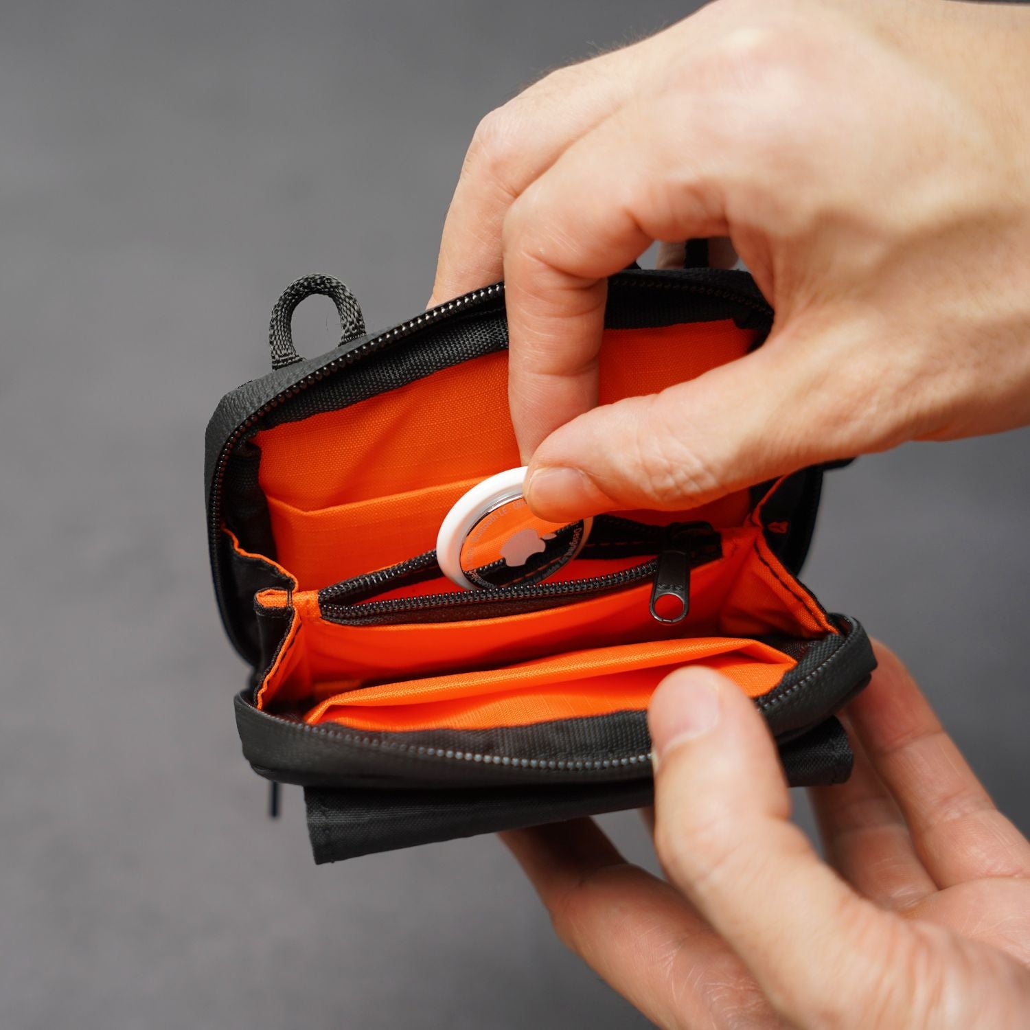 Person opening a small orange pouch with a black zipper on a gray background